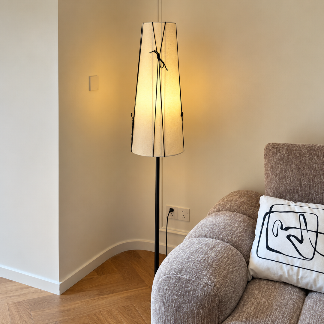 A modern floor lamp with a tapered off-white shade and thin black wire details, standing in a room corner next to a gray upholstered sofa with a white accent pillow.
