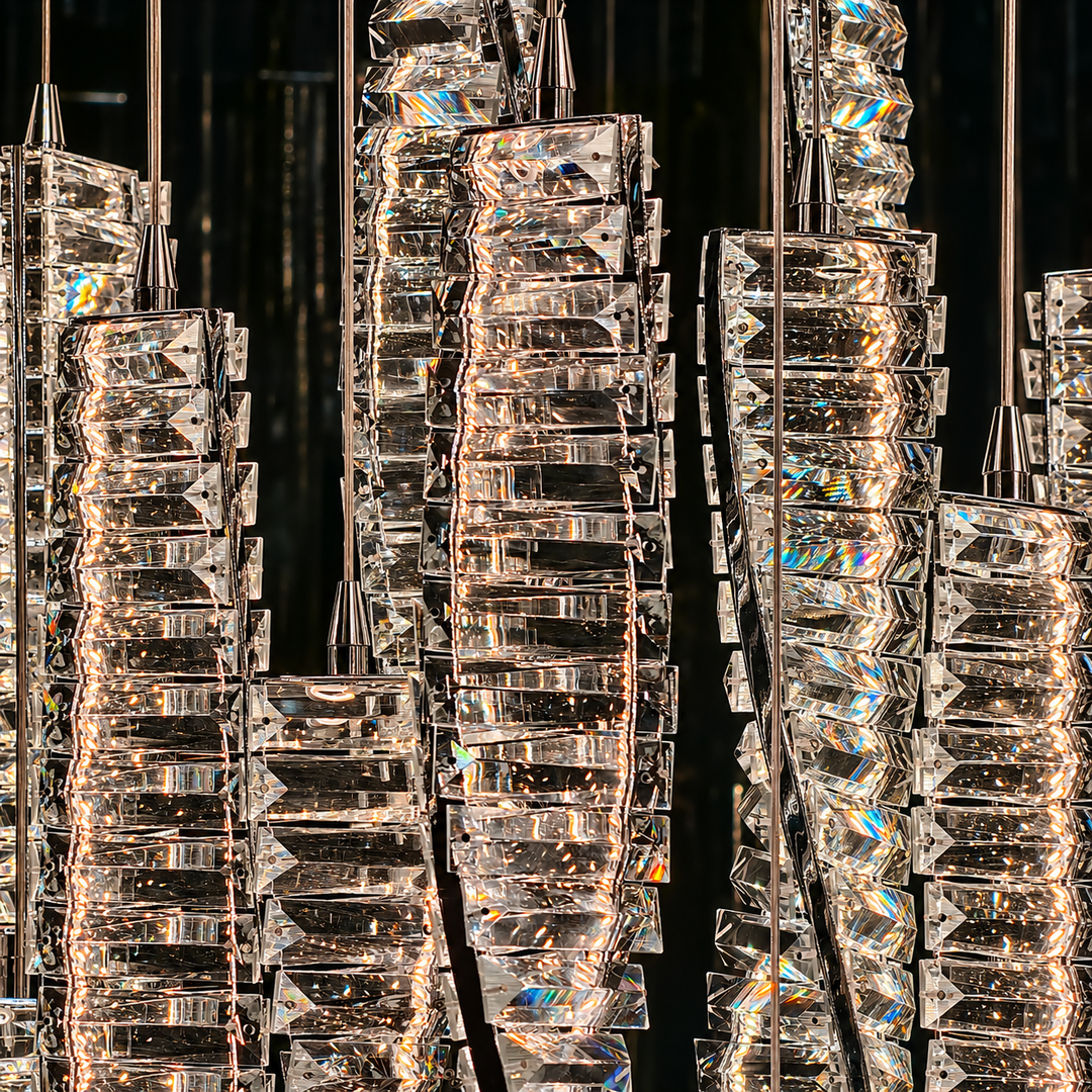 A close-up of a modern chandelier with multiple vertical clusters of clear faceted glass blocks and prisms, suspended by thin metal rods against a dark background. The transparent glass pieces catch and reflect warm light, creating bright rainbow reflections.