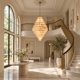 A grand, elegant interior entryway with a large tiered chandelier hanging in the center of a high-ceilinged hall with a sweeping staircase, marble floors, tall windows, and potted plants.