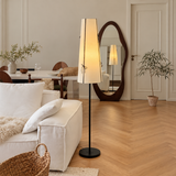 A tall floor lamp with a warm glowing white fabric shade and a black metal stand is placed in a bright, modern living room next to a white sofa. The room features light wood herringbone floors, a large organic-shaped wooden mirror, and a dining table in the background.
