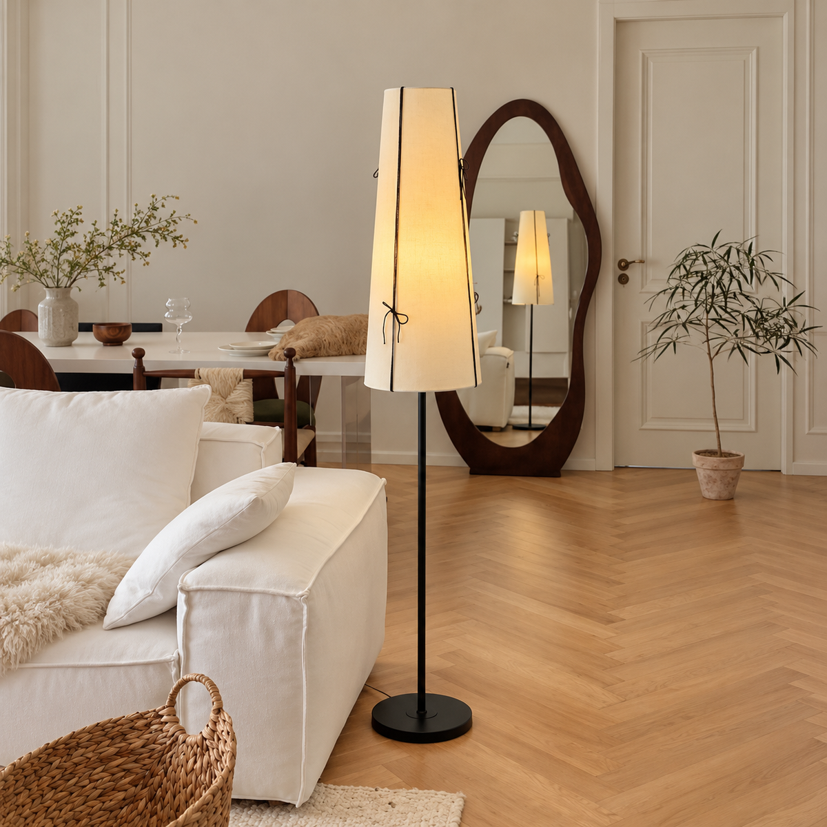 A tall floor lamp with a warm glowing white fabric shade and a black metal stand is placed in a bright, modern living room next to a white sofa. The room features light wood herringbone floors, a large organic-shaped wooden mirror, and a dining table in the background.