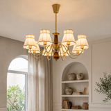 A multi-arm brass chandelier with cream fabric drum shades hangs in a warm, elegant room with built-in shelves, arched windows, and neutral-toned decor.