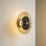 Circular wall sconce with an amber outer disc, smoky dark inner disc, and horizontal metal bar casts warm light on a cream wall.