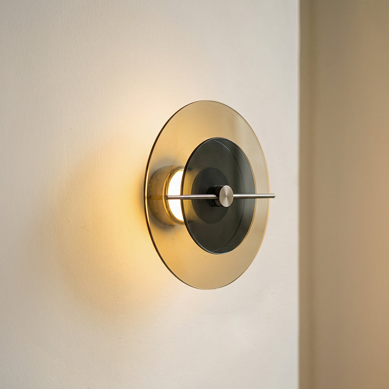 Circular wall sconce with an amber outer disc, smoky dark inner disc, and horizontal metal bar casts warm light on a cream wall.