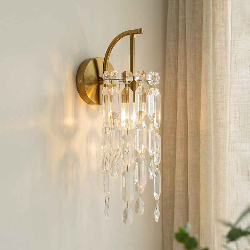 A wall-mounted sconce with a brushed gold metal frame and clear hanging crystal prisms, shown lit on an off-white wall beside beige curtains.