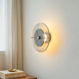Glass wall sconce casts a warm glow on a pale wall beside curtains and above a table with stacked books.