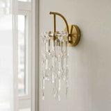 A wall-mounted brass sconce with clear hanging crystal prisms, shown installed on a light-colored wall next to a window.