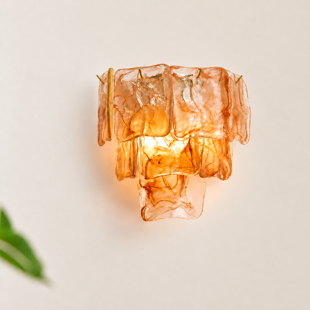 A sculptural wall sconce made from stacked translucent amber-orange glass blocks, glowing from within against a warm off-white background.