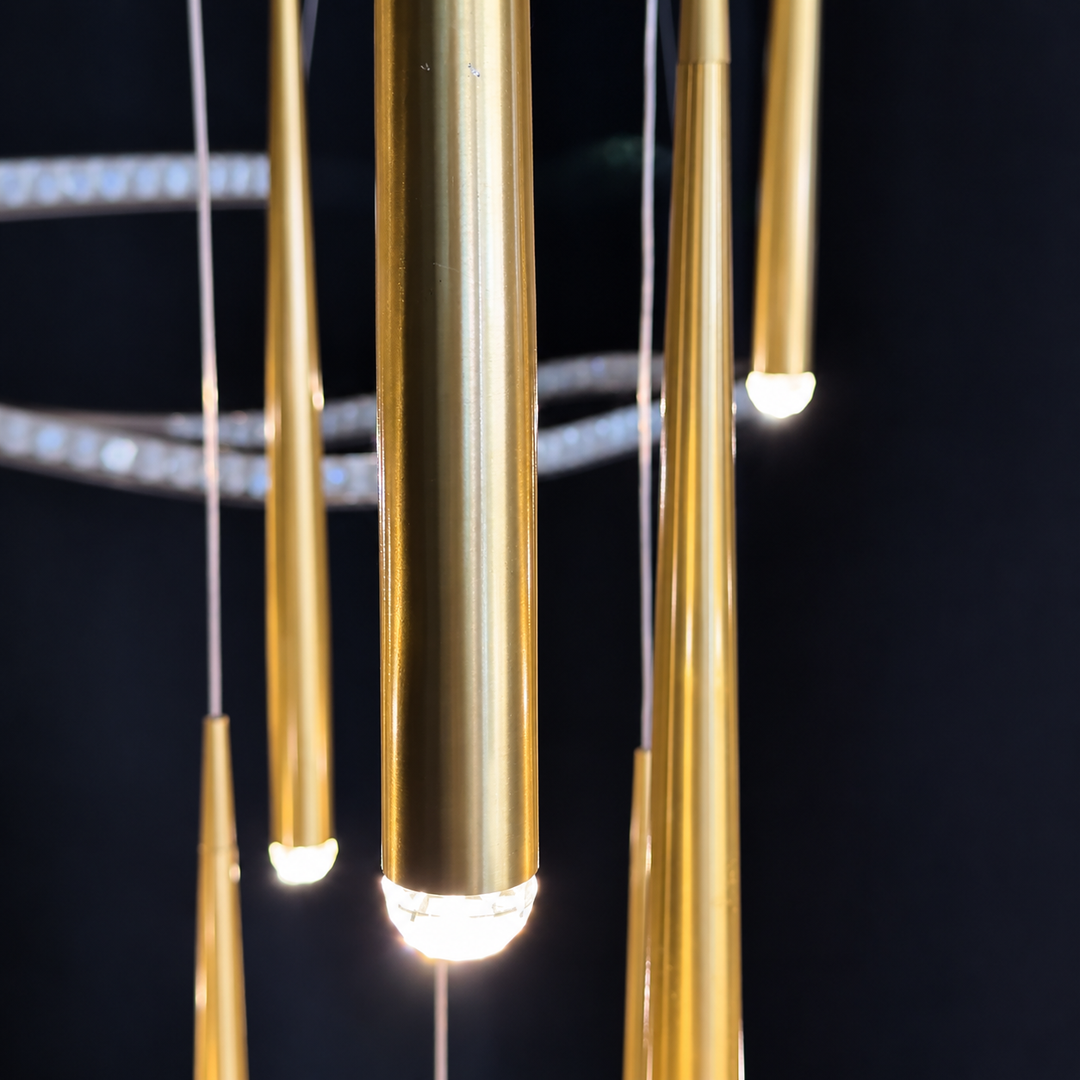 A close-up of a modern chandelier with brushed gold teardrop-shaped pendant lights and a circular ring of clear crystal-like beads, shown against a dark background.
