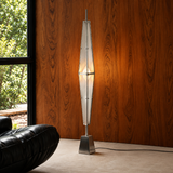A modern floor lamp with a tall, tapered white ribbed shade and a square metallic base, standing in a room with a wood-paneled wall and a window.