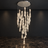 A modern multi-light chandelier with numerous illuminated crystal-like drop pendants suspended from a round silver ceiling canopy, shown hanging in a dark interior with a marble floor.