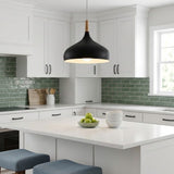 A modern kitchen scene with a black dome pendant light hanging over a white island. The kitchen features white cabinets, a green subway tile backsplash, and blue-gray bar stools at the front of the island.