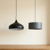 Two modern pendant lights hanging against a warm beige wall above a wooden table. The left light has a black dome shade with a light wood neck accent, and the right light has a matte dark gray cylindrical drum shade.