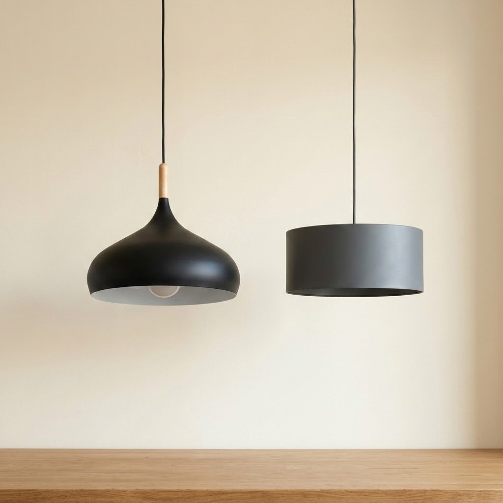 Two modern pendant lights hanging against a warm beige wall above a wooden table. The left light has a black dome shade with a light wood neck accent, and the right light has a matte dark gray cylindrical drum shade.