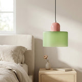 A light green glass chandelier with a glossy pink top hangs above a bedside table with a vase and book.