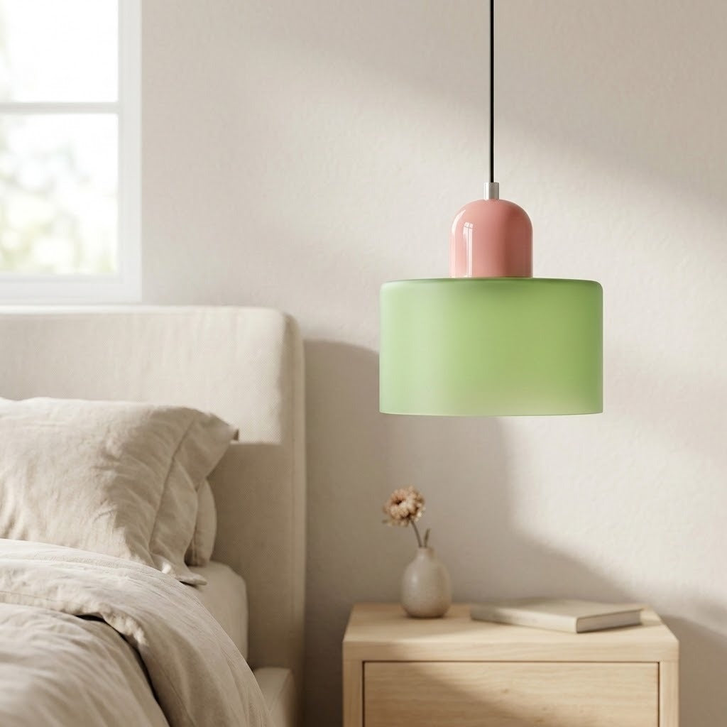 A light green glass chandelier with a glossy pink top hangs above a bedside table with a vase and book.