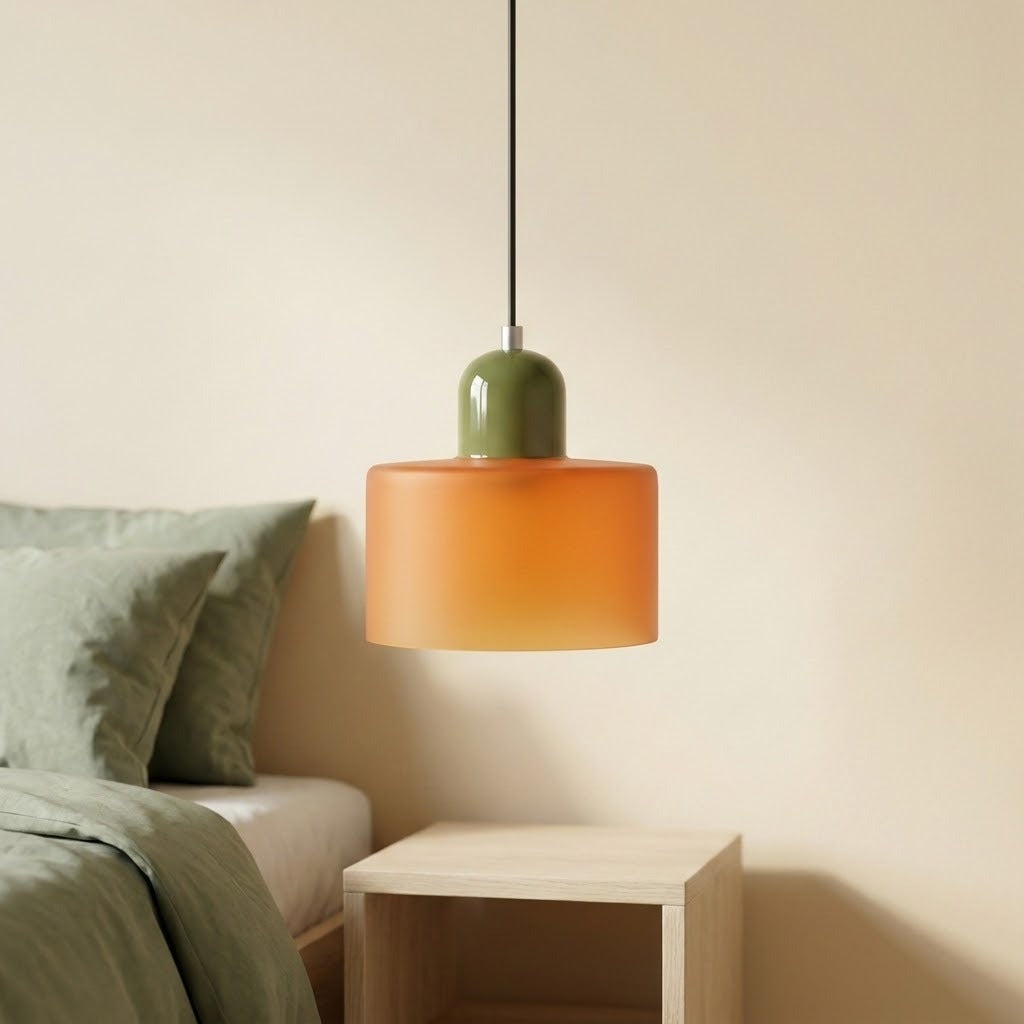 An orange glass chandelier with an olive green cap hangs above a pale wooden nightstand beside a bed with green bedding.