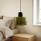 An olive green glass chandelier with a glossy dark cap hangs above a wooden nightstand beside a bed and window.
