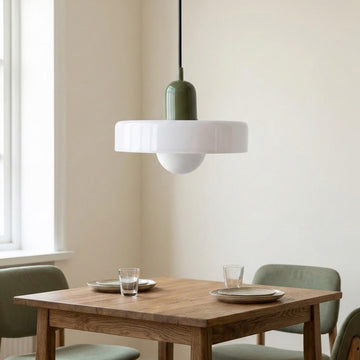 A modern pendant light with a white flared shade and an olive green cylindrical neck hangs above a wooden dining table set with plates and glasses in a bright room.