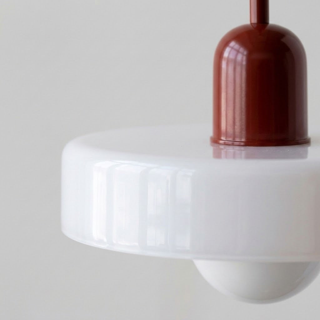 Close-up of a modern pendant light with a glossy white circular shade and a dark reddish-brown central cap and cord, set against a plain light gray background.