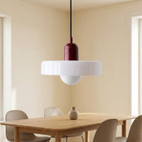 A modern pendant light with a glossy white circular shade and a dark burgundy cylindrical top, hanging in a dining room above a wooden table with chairs.