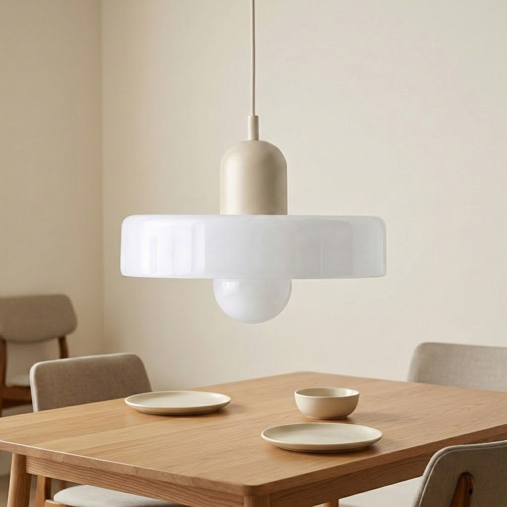 A modern pendant light with a wide white frosted glass shade and a warm beige cylindrical top, hanging over a wooden dining table in a minimalist room.