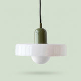 A modern pendant light with a wide, flared white glass shade, a glossy olive green central body, and a black hanging cord, shown against a solid pale green background.