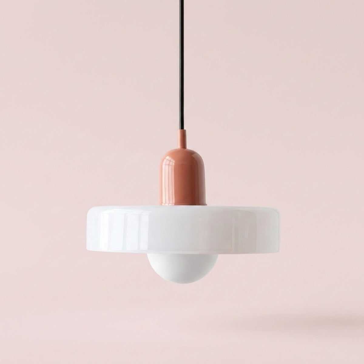 A modern pendant light with a wide white circular shade, a white dome bulb holder, and a terracotta-colored central cap on a black cord, set against a soft pink background.
