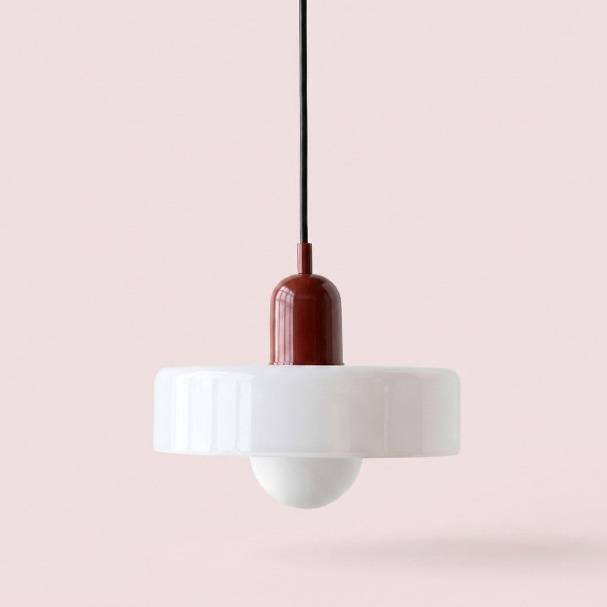 A modern pendant light with a wide white flared shade, a white globe bulb, and a dark reddish-brown central connector on a black cord, set against a solid pale pink background.