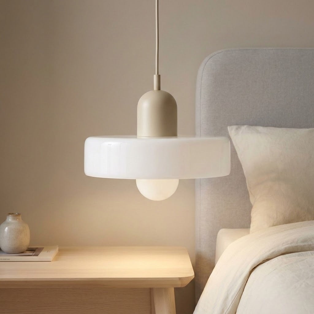 A modern pendant light with a matte beige top and a wide white frosted glass shade, hanging in a softly lit bedroom next to a bed with a light gray headboard.
