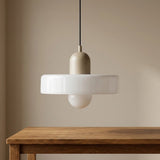 A modern pendant light with a matte beige cylindrical shade, a wide white circular glass shade, and a small white globe bulb underneath, hanging above a wooden table against a beige wall.