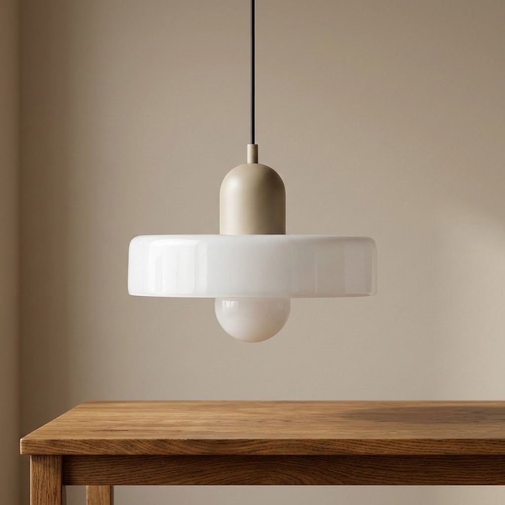 A modern pendant light with a matte beige cylindrical shade, a wide white circular glass shade, and a small white globe bulb underneath, hanging above a wooden table against a beige wall.