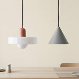 Two modern pendant lights hanging above a light wood table against a warm beige wall. The left light has a white circular shade, a terracotta-colored cylindrical bulb, and a white globe bulb.