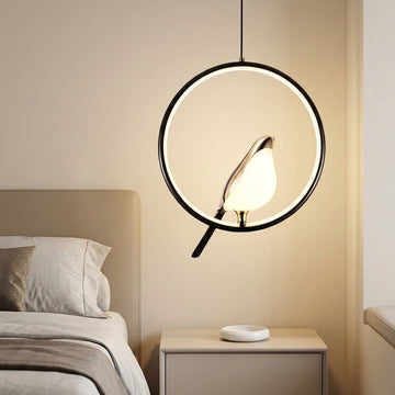 A modern circular pendant light with a black metal frame and a glowing bird-shaped bulb, hanging in a softly lit bedroom above a bedside table.