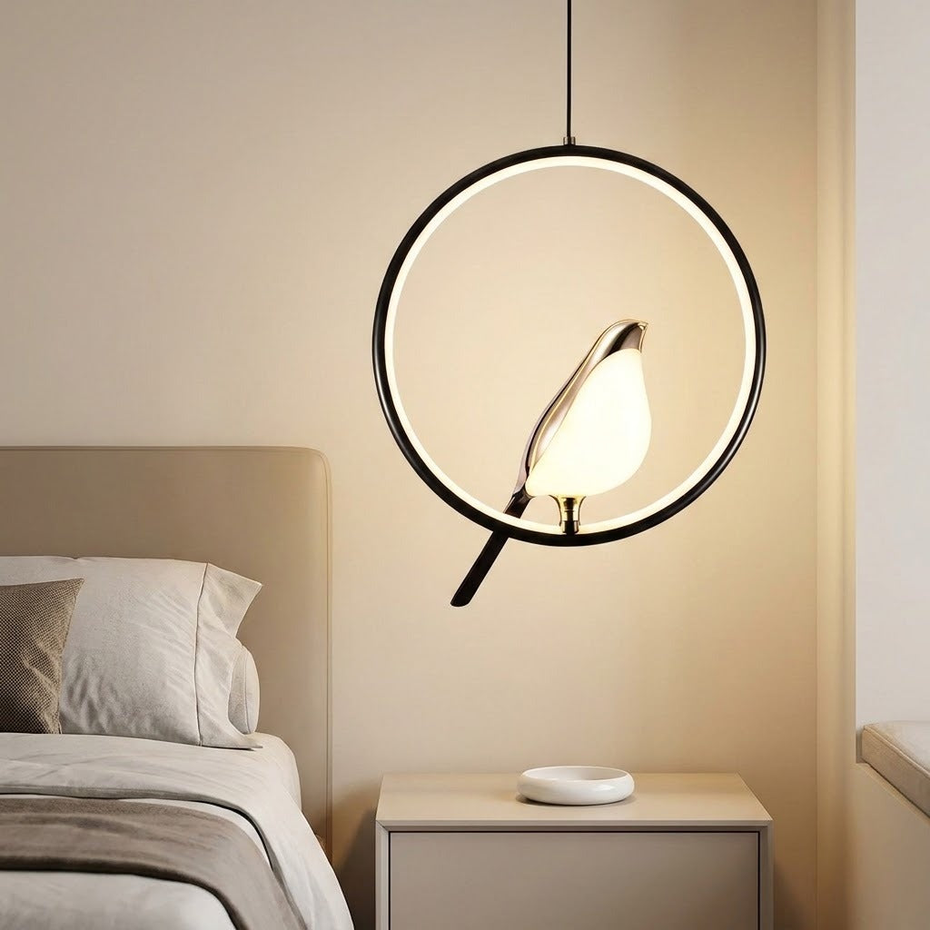 A modern circular pendant light with a black metal frame and a glowing bird-shaped bulb, hanging in a softly lit bedroom above a bedside table.