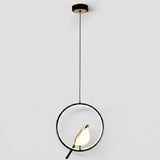 Modern pendant light with a black circular ring frame, a black cord, a black ceiling canopy, and a small gold-toned bird-shaped bulb holder containing a glowing warm white bulb, shown against a light gray background.