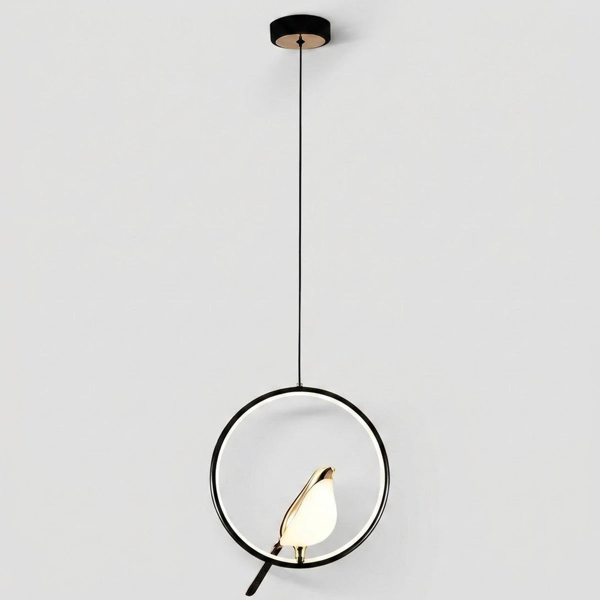Modern pendant light with a black circular ring frame, a black cord, a black ceiling canopy, and a small gold-toned bird-shaped bulb holder containing a glowing warm white bulb, shown against a light gray background.