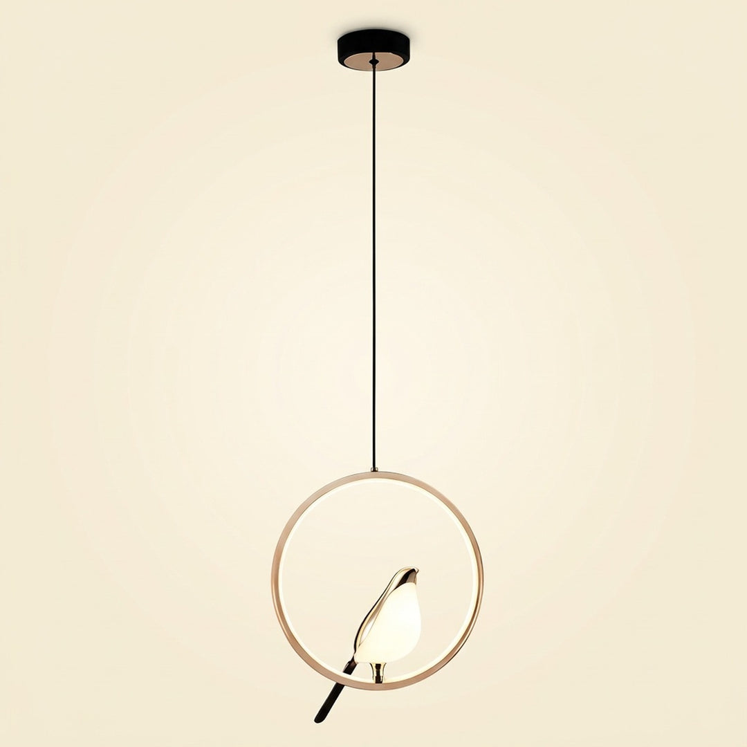 A modern pendant light with a thin circular gold-toned frame, a black hanging cord, a black ceiling canopy, and a small bird-shaped bulb or light element inside the ring, shown against a plain warm off-white background.