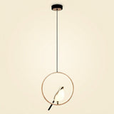 A modern pendant light with a thin circular gold-toned frame, a black hanging cord, a black ceiling canopy, and a small bird-shaped bulb or light element inside the ring, shown against a plain warm off-white background.