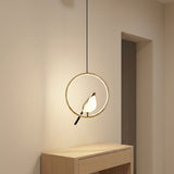 A modern pendant light with a circular gold frame and a bird-shaped bulb, hanging above a light wood console table in a minimalist room.