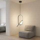 A modern oval pendant light with a black metal frame and a warm glowing white glass shade, hanging in a minimalist beige room next to a gray upholstered lounge chair.