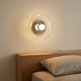 Clear glass wall sconce with a silver center casts warm light above a wooden headboard and neutral bedding.