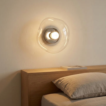 Clear glass wall sconce with a silver center casts warm light above a wooden headboard and neutral bedding.