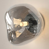 An organic-shaped wall sconce with a transparent smoky glass shade and a central cream-colored bulb holder, mounted on a light beige wall.
