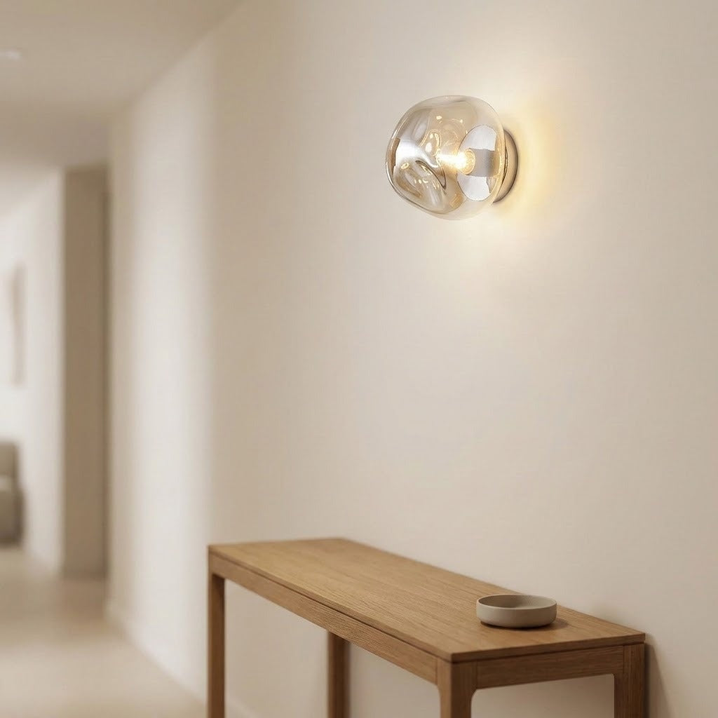 A modern spherical wall sconce with a clear glass globe and warm glowing bulb, mounted on a cream-colored wall above a light wood console table.