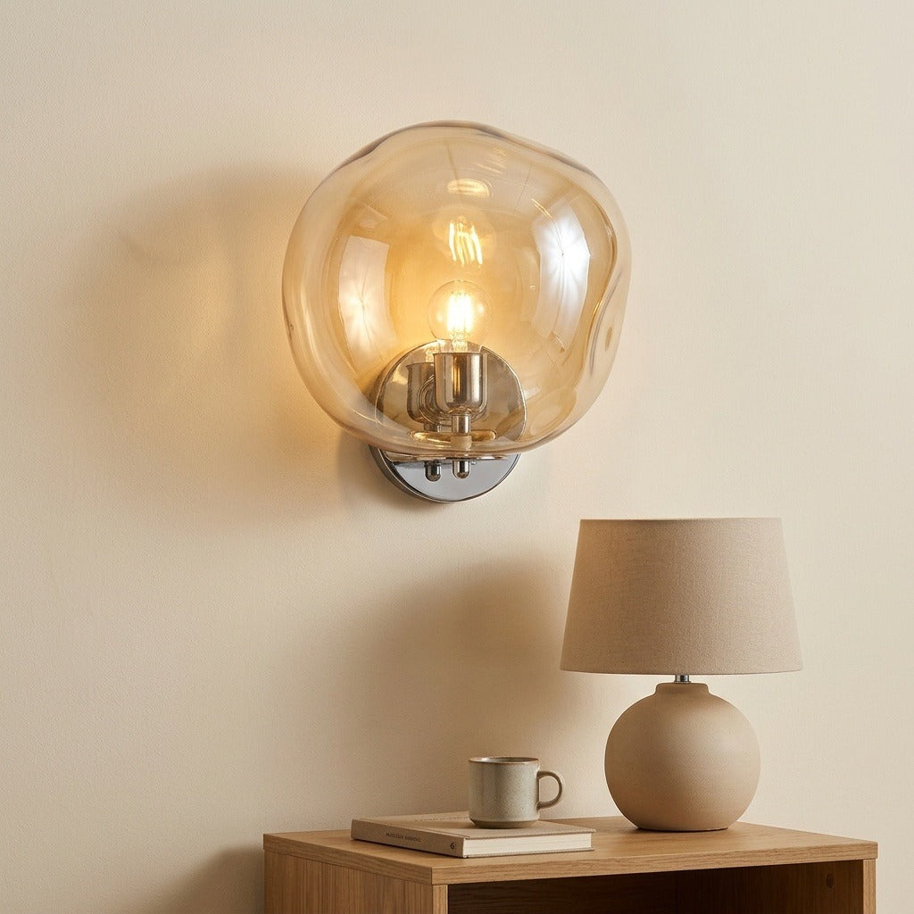 A modern wall-mounted sconce with a large, organic-shaped translucent amber glass shade and a silver metal base, shown lit on a beige wall above a wooden side table with a lamp and a mug.