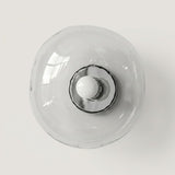 Clear glass wall sconce with an irregular rounded shade, polished silver center, and white bulb against a light neutral background.