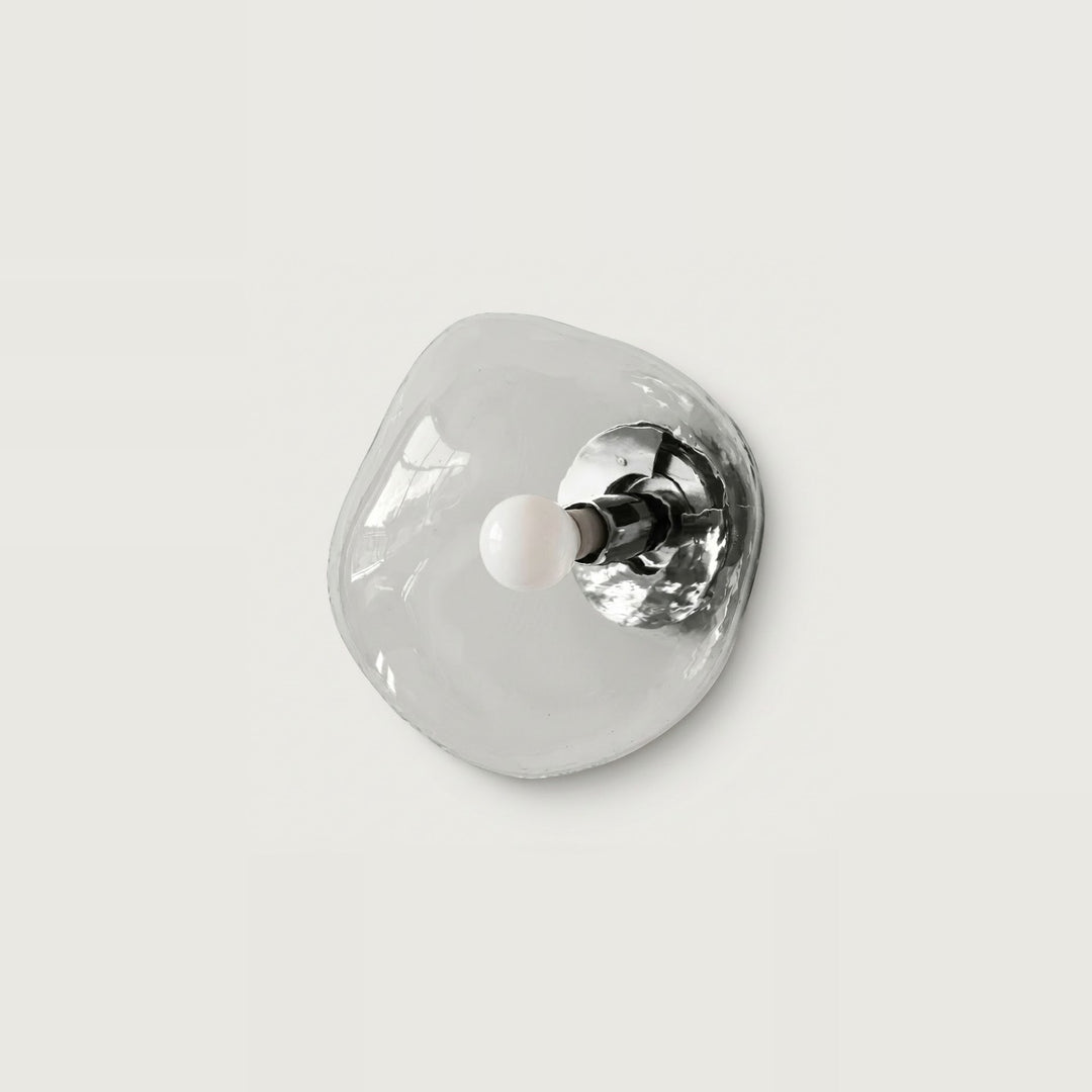 An organic-shaped, translucent clear glass wall sconce with a polished silver metal backplate and a small white spherical bulb, shown against a solid light gray background.