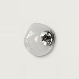 An organic-shaped, translucent clear glass wall sconce with a polished silver metal backplate and a small white spherical bulb, shown against a solid light gray background.