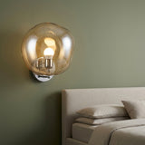 A modern wall-mounted sconce with a large, organic-shaped translucent amber glass globe and a warm glowing bulb, mounted on a sage green wall above a bed.